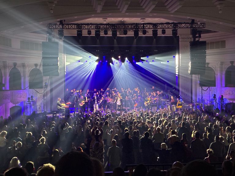 The Cat Empire performs live on a stage illuminated by bright blue spotlights before a large, cheering crowd inside an ornate theater hall.